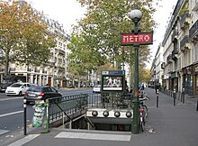 Frontale Farbfotografie eines Metro-Eingangs mit grünem Geländer und grüner Laterne, an der das rote Schild mit weißer Aufschrift „Metro“ hängt. An der Decke hängen vier runde Lampen und im Hintergrund ist ein Kasten mit der Aufschrift „M8 Saint-Sébastien Froissart“ und einem Plakat für Uhren. Im Hintergrund ist eine Allee mit alten Häuserfassaden.