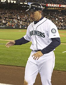 An olive-skinned man in a white baseball uniform with "Mariners" across the chest and a dark baseball cap