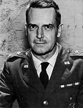 Man with dark hair and moustache in a dress uniform, consisting of a military suit and tie, sitting at a table, with two stars on each lapel indicating his rank, in front of a world map on a wall.