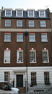 A terrace house with four floors and an attic. It is red brick, with a slate roof, and the ground floor rendered in imitation of stone and painted white. Each upper floor has four sash windows, divided into small panes. The door, with a canopy over it, occupies the place of the second window from the left on the ground floor.