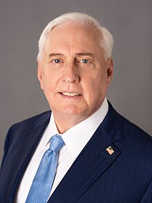 A portrait of Macgregor smiling in front of a grey background. He wears a navy blue suit jacket with an American flag lapel pin, a white shirt, and a light blue necktie.