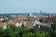 Farbige Panoramaansicht einer Stadt mit alten Gebäuden und Bäumen im Vordergrund. Im rechten Hintergrund sind Wolkenkratzer und links hinter einem Wald sind moderne Häuser.