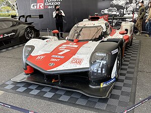 A front view of a red and white Toyota GR010 Hybrid on display at a car show