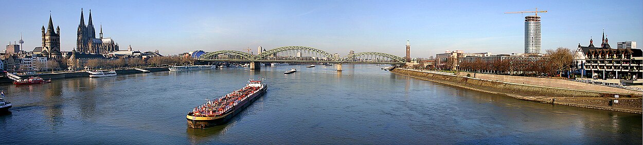 Panoramisch zicht op de Rijn bij Keulen vanaf de Deutzer Brücke. Geheel links in de verte de tv-toren, dan de kerk Groß St. Martin en de Dom, het blauwe dak van het musicalgebouw, de brug Hohenzollernbrücke, aan de andere kant van de Rijn de toren van de Messe, en uiterst rechts een omstreden kantoortoren (toen nog in bouw).