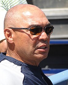 An older dark-skinned man wearing a blue-and-white shirt and sunglasses.