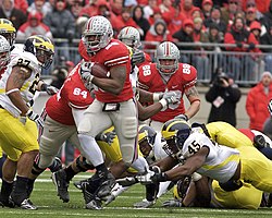 An American football player wearing scarlet and grey attempts to elude tacklers wearing Maize and Blue uniforms.