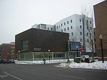 Modern building seen from across the street, cyclist out front, snow on the ground. Six story silver tower. Two story entrance. First floor is glass, second floor is high-ceilinged, dark solid with window in front.