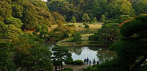 A panoramic view of the Rikugi-en from the Fujishiro-toge hill vantage point. Green trees surround a serene lake.