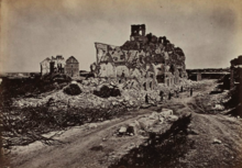 Photographie du fort d'Issy, détruit par la guerre. Il est situé au bord d'une route, sur une butte de gravats entourée d'arbres morts. On ne distingue du fort que des morceaux de façade, aucune d'entre elles n'ayant été épargnée par les bombardements.