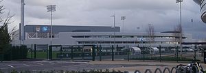 Photo du bâtiment gris où se situe la City Football Academy. Le ciel est nuageux.