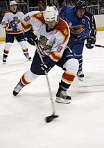 An ice hockey player is clearing the puck on his backhand, he has his head down looking at the puck. He is wearing a "traditional" Panthers white jersey. In the background a teammate is yelling to him while an opposing player, in a blue jersey with a stylized bird with a hockey stick crest logo, pressures him.