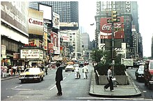 A view of Times Square in 1977 showing wast expanses of street, people crossing them in many locations and various billboards scattered on the buildings.