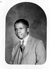 Black and white photo of a man in a suit facing forward towards a camera.