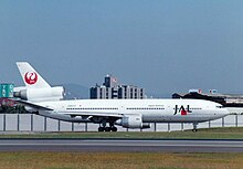 A McDonnell Douglas DC-10 aircraft taxiing on the tarmac, with a yellowish grass strip in the foreground and buildings and fence in the background