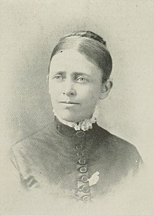 B&W portrait photograph of a woman with her hair in an up-do wearing a high-collared dark top with white fringe at the neckline.