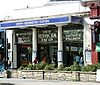 A entrance behind a raised bed of green plant and under a blue sign reading "SOUTH KENSINGTON STATION" leads into an arcade of shops. Above the entrance there is a glass panel with white lettering reading "METROPOLITAN AND DISTRICT RAILWAYS" and "SOUTH KENSINGTON STATION".