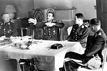 Black-and-white photograph of four men wearing uniforms sitting on wooden chairs around a table in a living room. An older man is sitting at the head of the table on the left. Two younger men are sitting along the table's side, with their backs toward a tiled fireplace on the room's far wall. A fourth man is sitting at the head of the table on the right, leaning back, his left leg folded over his right. The table is covered by a white table cloth. The right-hand side of the table is empty, except for a large dark ashtray, a spoon and an empty glass; the left and centre of the table is covered by an assortment of empty plates, coffee cups and other dishes; there is also a bowl of fruit. The second man from the left has his hands raised, palms facing the camera, and the fingers of both hands pointing to the left of the image. The heads of the three other men are turned toward him.