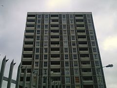 Eleven storeys of a gray residential tower, with a spiked fence and a lamp in the foreground.