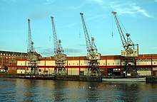 A long two-storey building with 4 cranes in front on the quayside. Two tugboats are moored at the quay.