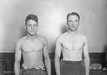 Deux hommes torse nu, côte à côte, devant un mur, les bras le long du corps.