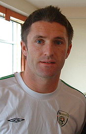 Head and torso of a young man. He has short brown hair with a quiff at the front, blue eyes, and is wearing a white t-shirt.