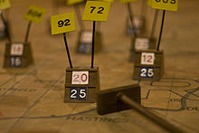 A wooden marker on a map, surrounded by several others. Below and to the right is a cue with a wooden slab on the end, used to move the markers.