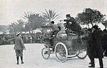 Trois hommes debout dans une voiture haute devant une foule contenue.