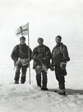 Three men stand around a flag planted in the snow.