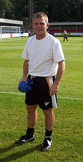 Um homem de cabelos loiros de 40 anos, vestindo uma camiseta branca e um calção preto, fica perto da borda de um campo de futebol.