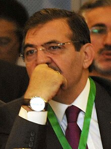 Colour photograph of İdris Güllüce aged 65 he is looking thoughtful with his hand resting against his chin. he is wearing a black suit with a maroon tie and a green lanyard. he is also wearing a white faced wristwatch and a pair of reading glasses.