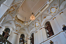 Statuary and cavettos of the Queen's Staircase, depicting Apollo, Artemis, cherubs, and the four classical elements. A glass chandelier (center) hangs from the ceiling at head height with the statues.