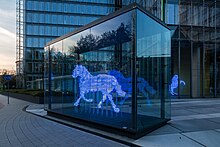 Photograph of a sculpture of a trotting horse. The sculpture was made using a series of horizontal bands. Each band is made from a glass tube that glows blue. The bands are stacked to indicate the form of a horse; the separation between the bands has about the same width as the glass tubing. The sculpture is inside a large glass box that has been placed on the plaza between some office buildings.