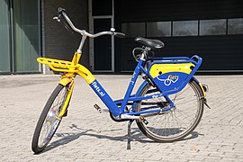 Farbfoto. Blau-gelbes E-Hollandrad, tiefer Einstieg, Beleuchtung, Gepäckträger vorne und hinten, Handbremse, Klingel. Auf dem Rahmen steht „OV-fiets.nl“. Städtischer Hintergrund.