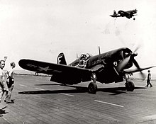 A Corsair on deck. A man stands nearby with fist upraised, giving a signal. There is another Corsair in the air above.