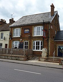 A photograph of a public house in a large red-bricked terrace house with a V-shaped slated roof, and an unpainted wooden picket fence and slab of ragstone at the front