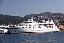 A white cruise ship in the water, docked along a coast with mountains in the background.