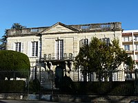 Bâtiment haut d'un étage et large de 5 hautes fenêtres, avec une rambarde sur son toit plat, précédée d'une terrasse et de sa grille de clôture.