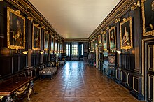 A photograph of a long dark room with high plain ceilings and gold-framed portraits lining the walls