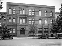 A rectangular, Victorian-style brick building with several trees and three cars from circa 1920 in front