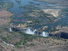 Farbige Vogelperspektive von mehreren Wasserfällen, die in eine Schlucht stürzen. Die Schlucht ist nach rechts offen. Der Fluss wird in der oberen Bildhälfte von Sandbuchten mit Bäumen unterbrochen. Unten ist eine trockene Landschaft mit wenigen Bäumen und einer Straße mit Lastkraftfahrzeugen.