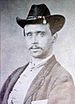 Head and shoulders of a white man with a thin mustache, wearing a cavalry hat, vest, and dark jacket.