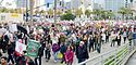 Marchers with signs walk down a street from right to left. Buildings and palm trees stand in the background.