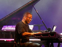 A Black man wearing a black t-shirt plays keyboard.