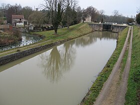 Vue vers l'amont depuis le pont de la Grande Rue : la Trézée s'apprête à rejoindre le canal. On ne voit pas le point de jonction, qui se trouve à quelques mètres derrière le pont ; on voit la Trézée qui se rapproche du canal et commence à le longer. La levée de terre entre le canal et la Trézée est renforcée sur les côtés par de larges pierres taillées et est surmontée d'un petit chemin piéton en terre battue et bordé d'une rambarde en métal côté Trézée (à gauche). Pour le canal, une écluse se trouve devant nous, à environ 90 en amont du pont d'où est prise la photo. Un chemin de halage borde le canal à droite, chemin goudronné d'ancien avec une bande d'herbe au milieu. La maison du garde-écluse est le seul bâtiment sur la gauche du canal. Il y a quelques maisons sur le côté droit du canal. Une seule maison se trouve le long de la Trézée en rive gauche (vu ici à droite du cours d'eau ; cette maison est entre la Trézée et la maison du garde-écluse) avec un petit pont sur la Trézée pour y accéder. Le cœur du village est hors de la photo sur la gauche, dans le prolongement de ce pont de la Rue Grande.