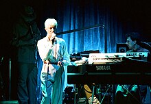 Photo d'un chanteur blanc aux cheveux clairs en costume tenant un micro près d'un joueur de synthétiseur, devant un rideau bleu