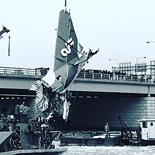 A severed airplane tail section hangs from a crane just above the water, guyed by crew on barges. A low, steel beam bridge with granite block piers stands behind, it's railing lined with onlookers.