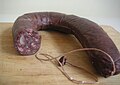 Sur une planche en bois, un morceau recourbé de boudin noir ; une extrémité est fermée par une ficelle, l'autre, tranchée net, montre les morceaux de graisses et la pâte de viande qu'enferme la peau de la charcuterie.