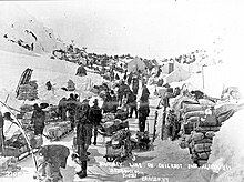 Peak of Chilkoot Pass in March-April 1898. Men wearing winter clothes with their supplies in the snow all of it surrounded by hill-sides.