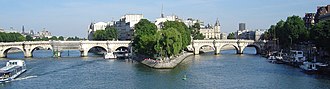 Frontale Farbfotografie eines Flusses, der in der Mitte von einer Insel geteilt wird. Die Insel verbindet beide Uferseiten mit zwei hellen Steinbrücken und ist mit vielen Gebäuden überfüllt. Der vordere Inselteil besteht aus breiten Bäumen und Menschen. An der linken Inselseite und am rechten Flussufer ankern Schiffe. Ein Ausflugsschiff fährt am linken Bildrand.