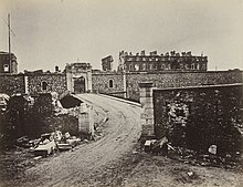 Photographie du fort de Vanvres, détruit par la guerre. Au premier plan, une route qui monte en sa direction, se finissant par un pont. Elle traverse un muret en partie détruit. Au deuxième plan, la muraille du fort et une entrée, situées derrière un fossé et peu endommagées. Derrière la muraille, on distingue quelques bâtiments dont une grande caserne à laquelle il ne reste plus que des pans de façades.